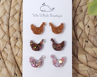 May include: A white card featuring six glittery chicken-shaped accessories. The chickens are in shades of brown, copper, and pink. The card has the text "Wee Whale Boutique" and a whale illustration.