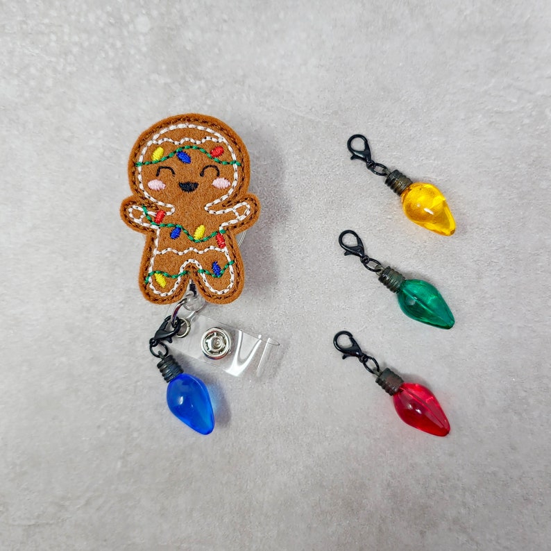 May include: A gingerbread man with colorful lights, a blue light bulb charm, and a retractable badge holder. Three additional light bulb charms are shown in red, green, and yellow.