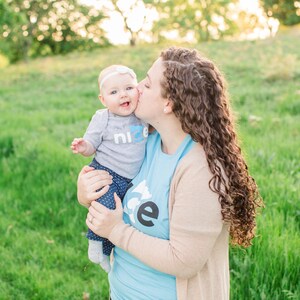 Puede incluir: Una mujer con cabello casta&ntilde;o rizado sostiene a una ni&ntilde;a peque&ntilde;a en sus brazos. La ni&ntilde;a lleva una camiseta gris con la palabra "nice" y una diadema blanca. La mujer lleva una camiseta azul con un dise&ntilde;o blanco y una chaqueta de punto beige. Ambas sonr&iacute;en y se miran.
