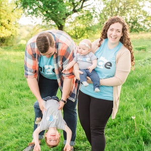 Puede incluir: Una familia de cuatro personas que llevan camisas azules con la palabra "nice" impresa en ellas. El padre lleva una camisa de cuadros sobre su camisa azul. La madre lleva un beb&eacute; en brazos. El beb&eacute; lleva una camisa gris con una diadema blanca. El ni&ntilde;o mayor est&aacute; boca abajo, sostenido por el padre. La familia est&aacute; de pie en un campo de hierba.
