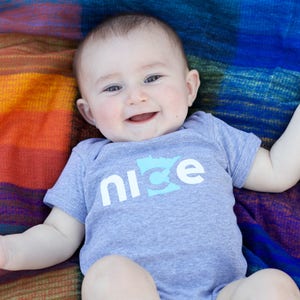 Puede incluir: Un beb&eacute; con un body gris con la palabra "nice" y un contorno del estado de Minnesota en azul claro. El beb&eacute; est&aacute; acostado sobre una manta colorida.