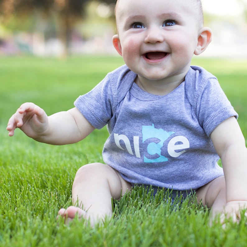 Puede incluir: Un beb&eacute; con una camiseta gris de manga corta con un contorno azul del estado de Minnesota y la palabra "nice" impresa en ella. El beb&eacute; est&aacute; sentado en la hierba verde y sonriendo.