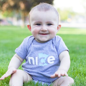 Puede incluir: Un beb&eacute; con una camiseta gris de manga corta con la palabra "nice" y un contorno del estado de Minnesota en azul. El beb&eacute; est&aacute; sentado en la hierba verde y sonriendo.