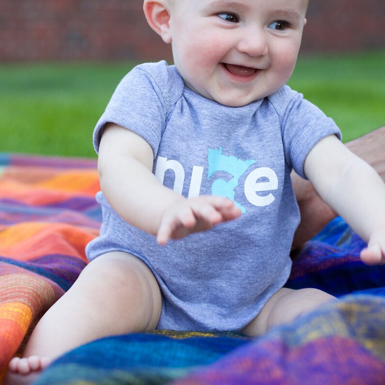 Puede incluir: Un beb&eacute; con un body gris con la palabra "nice" y un contorno azul del estado de Minnesota. El beb&eacute; est&aacute; sentado en una manta colorida.
