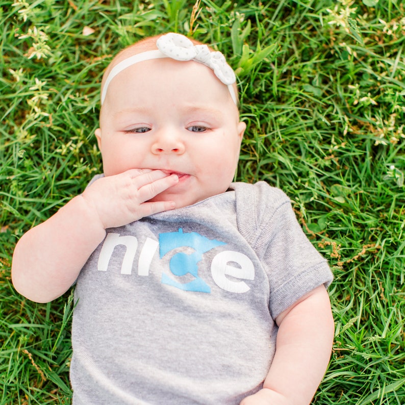 Puede incluir: Un beb&eacute; con una camiseta gris que dice "nice" y un contorno azul de un estado. El beb&eacute; est&aacute; tumbado de espaldas en la hierba y lleva una diadema blanca con un lazo.
