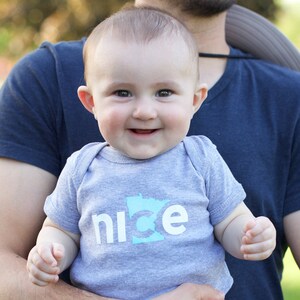Puede incluir: Un beb&eacute; con un mono gris con un contorno azul del estado de Minnesota y la palabra "nice" impresa en blanco.