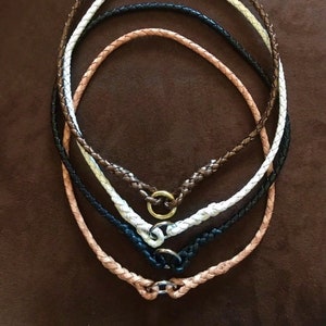 Puede incluir: Cuatro collares de cuero trenzado en diferentes colores, incluyendo marrón, blanco, negro y beige, con un cierre de anillo de plata.