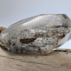 May include: A clear quartz crystal with inclusions of black and brown minerals. The crystal is faceted and has a geometric shape. It is resting on a piece of driftwood.