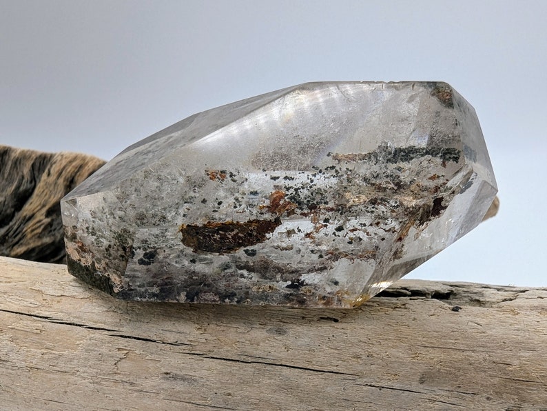 May include: A clear quartz crystal with inclusions of black and brown minerals. The crystal is faceted and has a geometric shape. It is resting on a piece of driftwood.