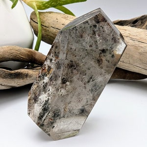 May include: A clear, faceted, gray crystal with black and brown inclusions. The crystal is shaped like a long, narrow rectangle with rounded edges.