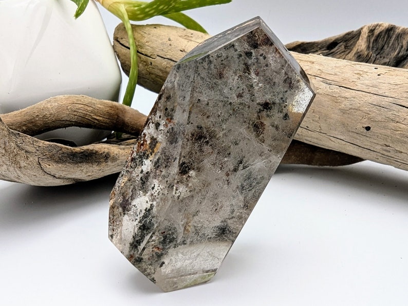 May include: A clear, faceted, gray crystal with black and brown inclusions. The crystal is shaped like a long, narrow rectangle with rounded edges.