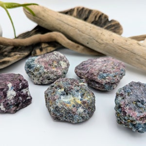 Op de afbeelding: Vijf ruwe, natuurlijke robijnkristallen in tinten roze, rood en paars. De kristallen zijn geclusterd op een wit oppervlak.