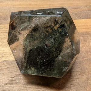 May include: A large, clear, faceted crystal with dark green inclusions. The crystal is shaped like a hexagon and has a smooth, polished surface.