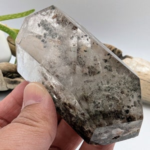 May include: A clear quartz crystal with black inclusions, shaped like a double-terminated point. The crystal is approximately 2 inches long and 1 inch wide.