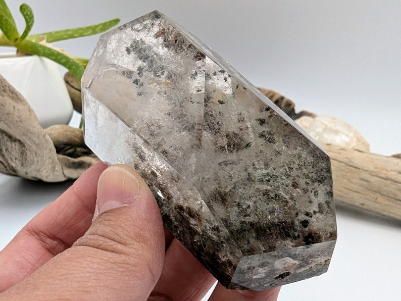 May include: A clear quartz crystal with black inclusions, shaped like a double-terminated point. The crystal is approximately 2 inches long and 1 inch wide.