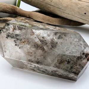 May include: A clear quartz crystal with inclusions of black and brown minerals. The crystal is faceted and has a pointed shape. It is approximately 3 inches long.