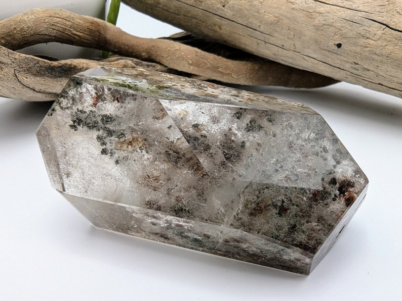 May include: A clear quartz crystal with inclusions of black and brown minerals. The crystal is faceted and has a pointed shape. It is approximately 3 inches long.