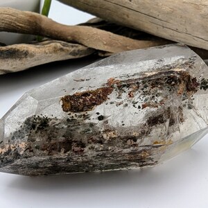 May include: A clear quartz crystal with inclusions of brown and black minerals. The crystal is faceted and has a pointed tip.