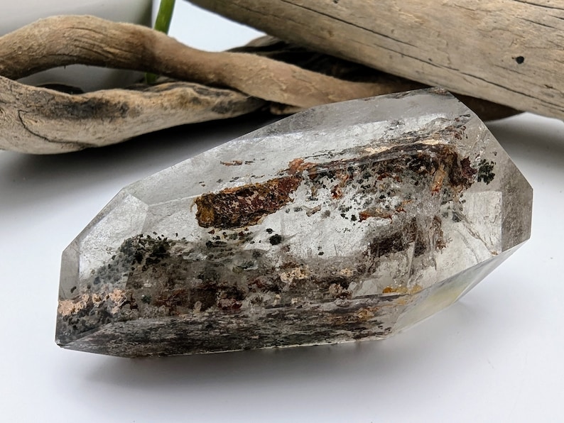 May include: A clear quartz crystal with inclusions of brown and black minerals. The crystal is faceted and has a pointed tip.