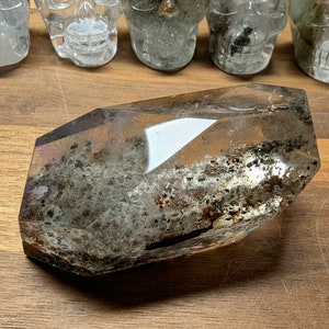 May include: A clear quartz crystal with a large, faceted point and inclusions of black and brown minerals. The crystal is lying on a wooden surface.
