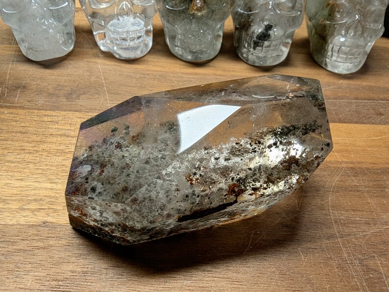 May include: A clear quartz crystal with a large, faceted point and inclusions of black and brown minerals. The crystal is lying on a wooden surface.