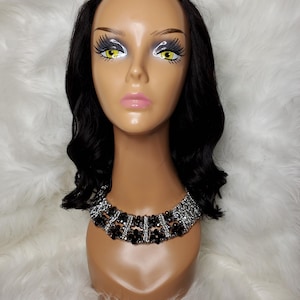 Puede incluir: Una cabeza de maniquí con cabello negro ondulado y un collar decorativo. El collar presenta cuentas negras y detalles plateados. El maniquí está sobre un fondo blanco y esponjoso.