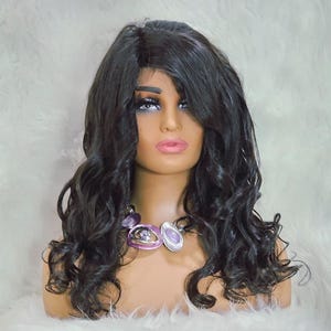 Puede incluir: Una cabeza de maniquí que muestra una peluca larga y ondulada de color negro con una raya a un lado. La peluca tiene rizos voluminosos y una línea de cabello de aspecto natural. El maniquí lleva un collar morado y plateado. El fondo es una superficie blanca y esponjosa.
