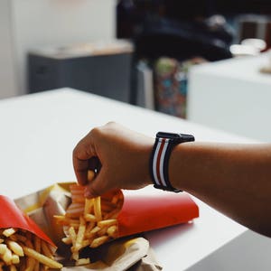 May include: A person's hand reaches for a container of french fries. The container is red with the McDonald's logo. The person is wearing a black and white striped watch band.