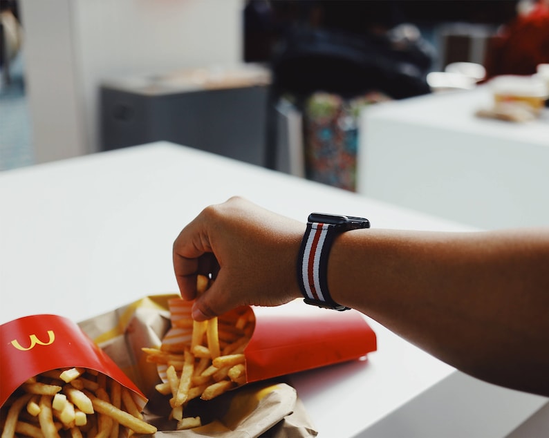 May include: A person's hand reaches for a container of french fries. The container is red with the McDonald's logo. The person is wearing a black and white striped watch band.