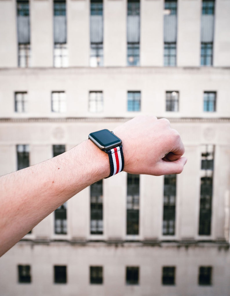 May include: A black Apple Watch with a red, white, and blue striped band. The watch is on a person's wrist.