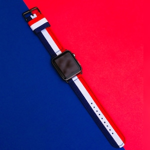 May include: A black smartwatch with a white, red, and blue striped nylon band. The watch is on a blue and red background.