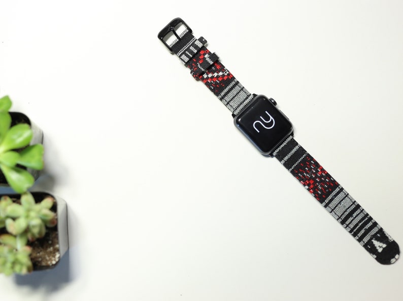 以下が含まれることがあります： 赤色のアクセントが付いた、黒と白の模様のApple Watchバンド。バンドは織り生地製で、黒いバックルが付いています。