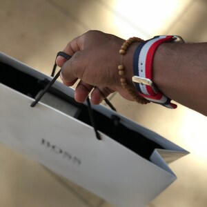 May include: A white shopping bag with the word "BOSS" printed on it. A person is holding the bag with their right hand. The person is wearing a red, white, and blue striped watch band with a wooden bead bracelet.