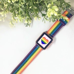 May include: A rose gold smartwatch with a rainbow-striped band. The watch face displays a rainbow heart. The band is approximately 1 inch wide. The watch is set against a white background with green foliage.