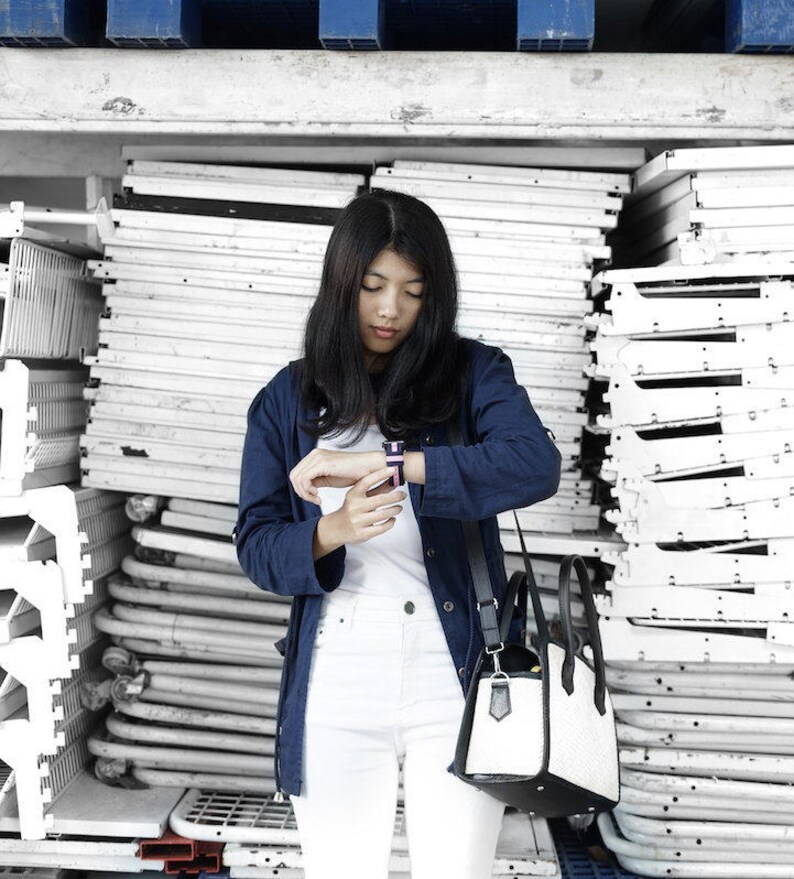 May include: A woman wearing a blue denim jacket, white pants, and a black and white handbag. She is checking the time on a smartwatch on her left wrist.