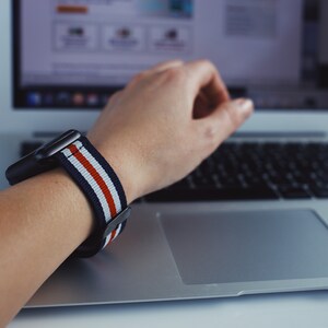 May include: A person wearing a black and white striped watch band with red stripes on a silver watch. The person's hand is resting on a silver laptop keyboard.