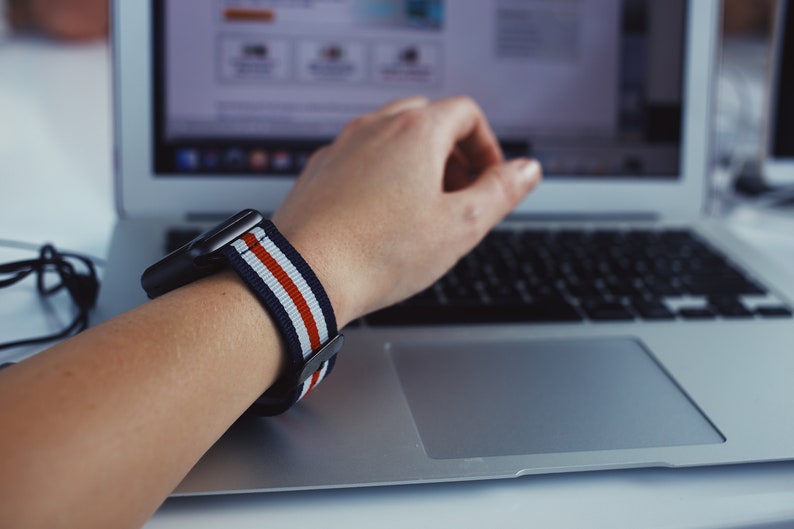 May include: A person wearing a black and white striped watch band with red stripes on a silver watch. The person's hand is resting on a silver laptop keyboard.