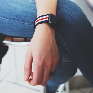 May include: A person wearing blue jeans with a watch with a red, white, and blue striped band.