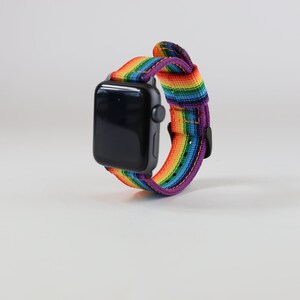 May include: A black smartwatch with a rainbow-striped band. The band features a vibrant spectrum of colors, including red, orange, yellow, green, blue, and purple. The watch is set against a plain white background.