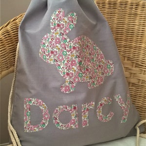 May include: Grey drawstring bag with a floral print bunny and the name "Darcy" in floral print letters.