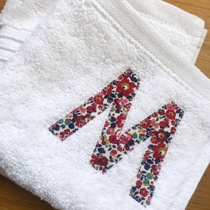 Może przedstawiać: Biały ręcznik z kwiatową aplikacją w kształcie litery "M". Litera wykonana jest z tkaniny z wzorem czerwonych, różowych i niebieskich kwiatów na białym tle. Ręcznik jest złożony i ma teksturowaną powierzchnię.