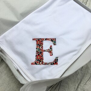 May include: A white baby blanket with a large letter E embroidered in the centre. The letter E is made up of a floral pattern in shades of red, blue, and green.