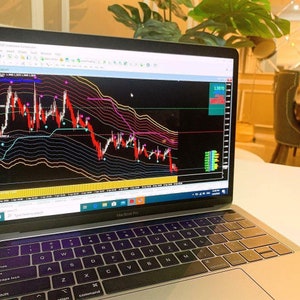 Puede incluir: Un ordenador portátil con una pantalla negra que muestra un gráfico de mercado de valores con líneas rojas y verdes. El gráfico muestra una tendencia a la baja. El portátil está abierto y el teclado es visible. El portátil está sobre una superficie blanca.