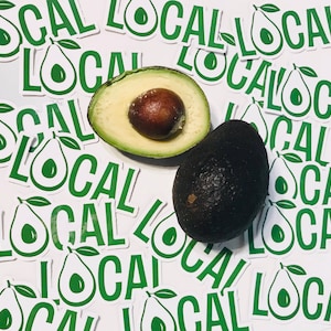 May include: A close-up of two avocados, one cut in half, surrounded by numerous stickers that say "Local" in green letters on a white background.