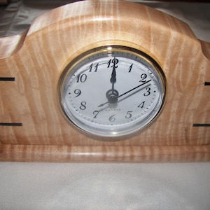 May include: A wooden clock with a light brown wood grain finish and black accents. The clock face is white with black numbers and hands. The clock is sitting on a white surface.