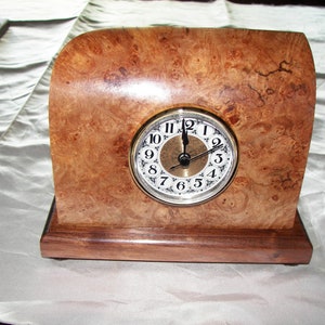 May include: A wooden clock with a burl wood case and a white face with black Roman numeral hour markers. The clock has a gold-colored trim and hands.