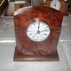 May include: A wooden clock with a white face and black Roman numeral hour markers. The clock is set to 12:00 and has a brown wood base.