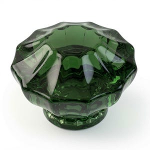 May include: A dark green glass cabinet knob featuring a faceted, multi-sided top with a scalloped edge and a circular base. The translucent material reflects light, highlighting the geometric, star-like pattern across the top surface.