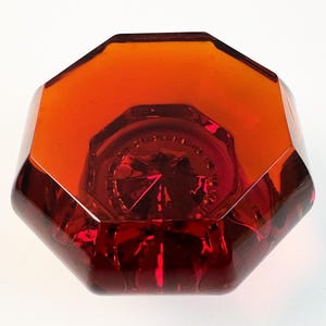 Puede incluir: Un pomo de puerta de cristal octogonal en un tono ámbar y rojo translúcido profundo. El pomo presenta un diseño geométrico facetado con un patrón central en forma de estrella visible a través del cristal, creando un aspecto multidimensional.