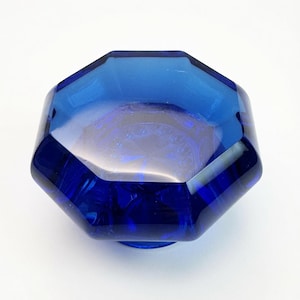 May include: A cobalt blue glass door knob with an octagonal shape and faceted edges. The surface is smooth and transparent, reflecting light across its geometric faces. The centre of the knob features a starburst pattern visible through the glass.